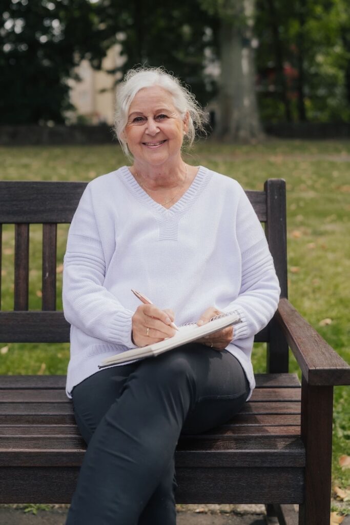 Martina Peters von erzähle uns dein Leben sitzt auf einer Parkbank und schaut lächelnd in die Kamera. Sie hält einen Schreibblock auf den Knieen in den sie Notizen schreibt.