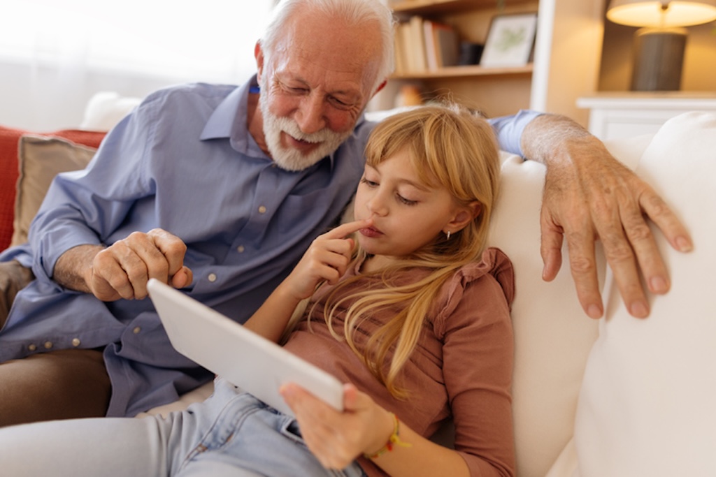 Herzerwärmende Szene einer Enkelin und ihres Großvaters, die zusammen auf einem gemütlichen Sofa sitzen und auf ein digitales Tablet schauen. Beide sind vertieft. Die warme, einladende Wohnzimmeratmosphäre verstärkt das Gefühl der Zweisamkeit, während sie gemeinsam Spaß daran haben, etwas Neues zu lernen oder zu entdecken.